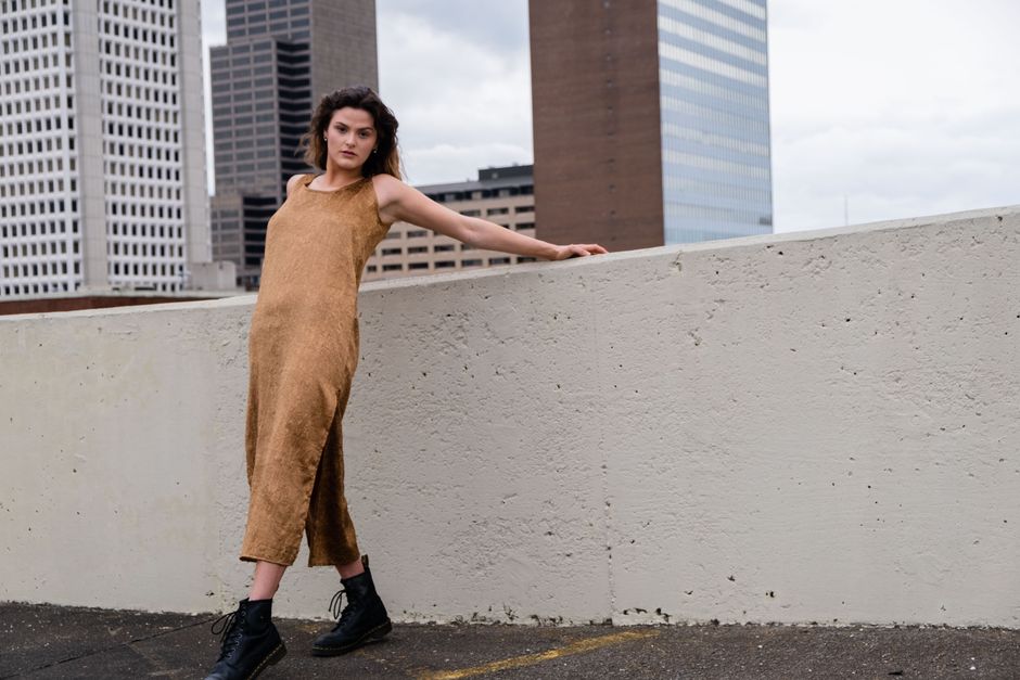 Woman in a brown dress and boots posing on a city rooftop with tall buildings in the background.
