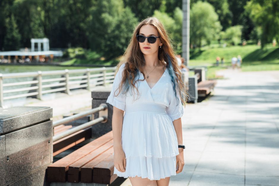 A young woman in a white dress and sunglasses enjoys a sunny day outdoors.