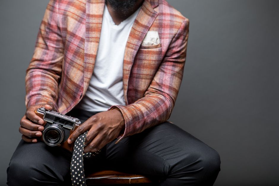 A fashionable man in a plaid blazer holds a vintage camera, seated indoors.