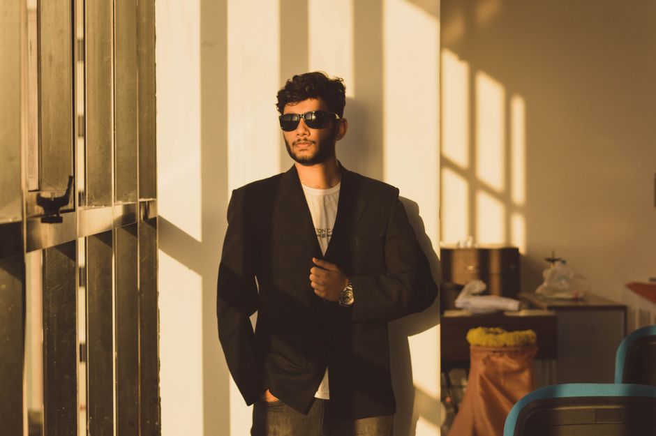 A fashionable man wearing sunglasses and a suit leans confidently against a sunlit wall.