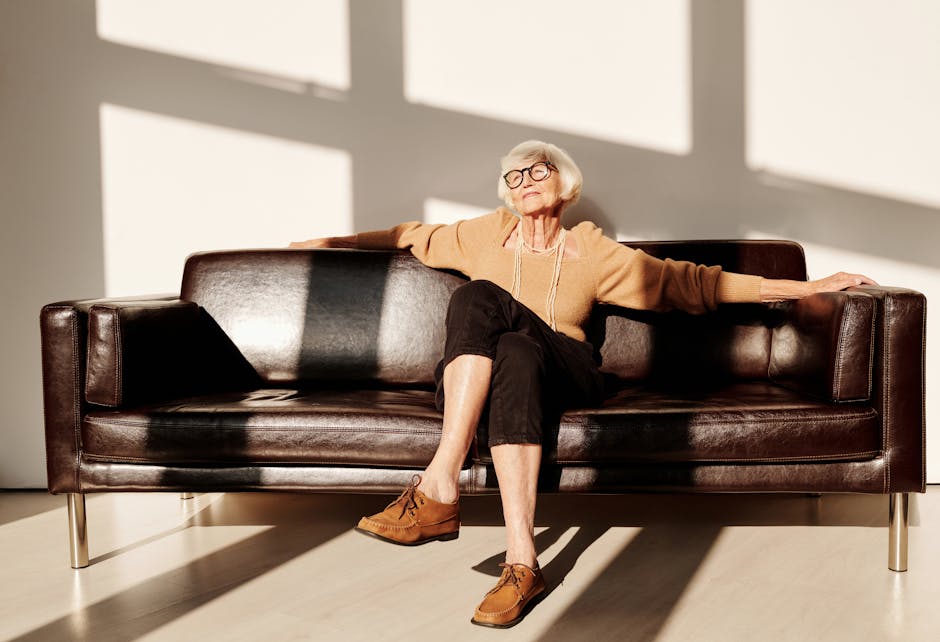 Elderly woman with gray hair and eyeglasses relaxing on a leather sofa in a bright room.