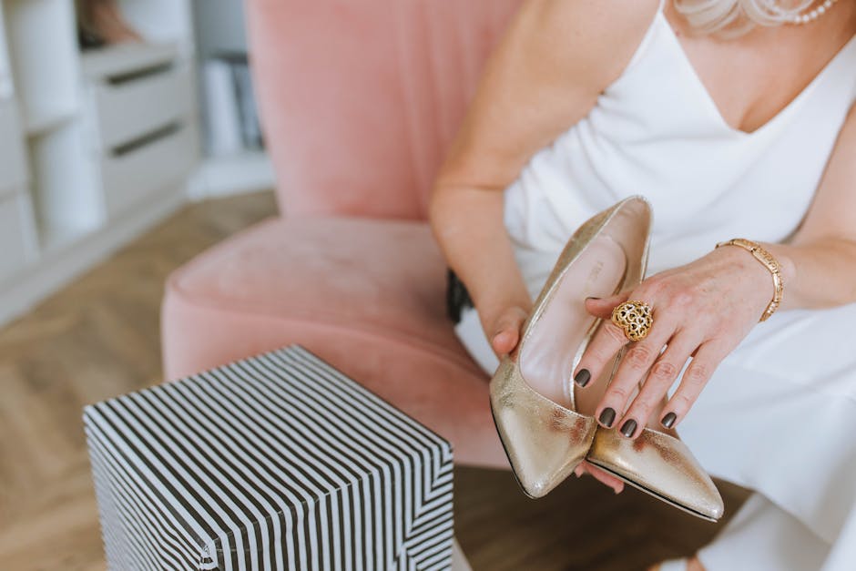 Stylish woman with jewelry and manicured nails holding elegant shoes near a gift box.