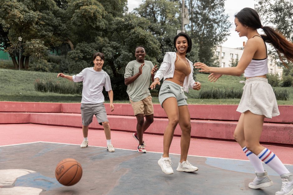 Group of excited young multiracial friends in activewear playing basketball together and laughing happily while gathering on sports ground in lush summer park