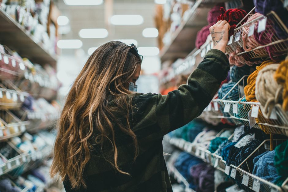 A woman wearing a mask browsing colorful yarn in a craft store aisle.