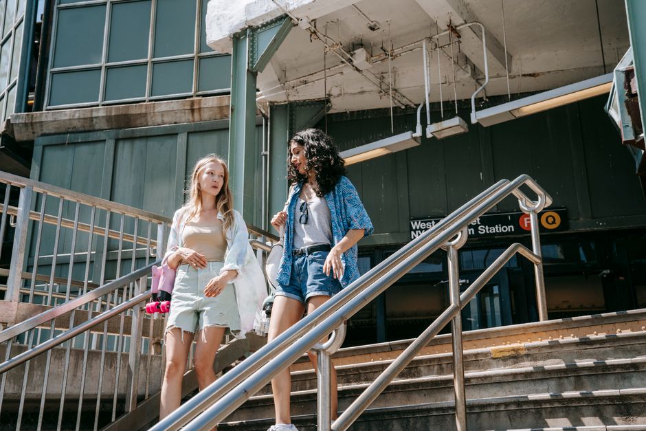 Two women descend from a city subway station under bright daylight, depicting urban life.