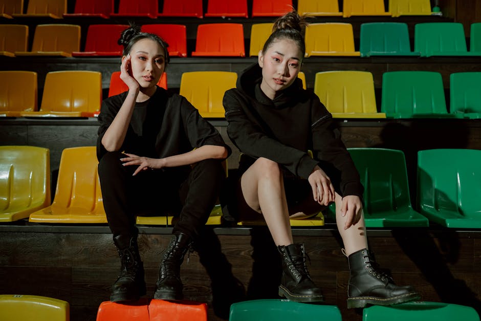 Two women in black outfits sit on colorful stadium seats indoors, striking a chilling pose.