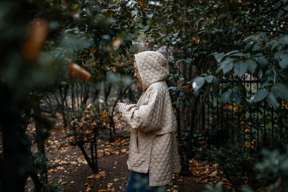 A woman in a beige quilted jacket strolls through an autumn park surrounded by lush greenery and fallen leaves.