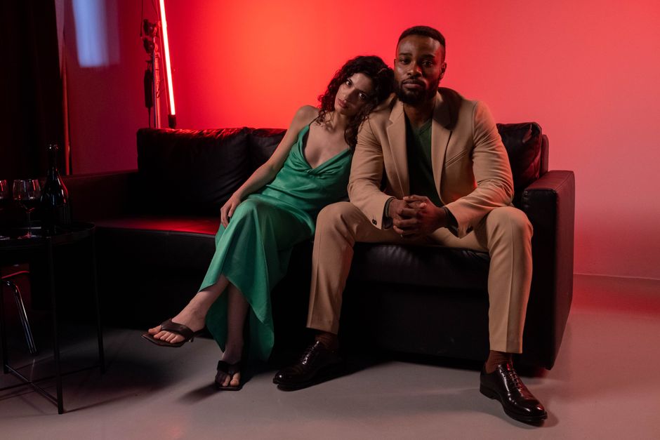 Couple sitting on leather couch in stylish outfits, dramatic lighting.