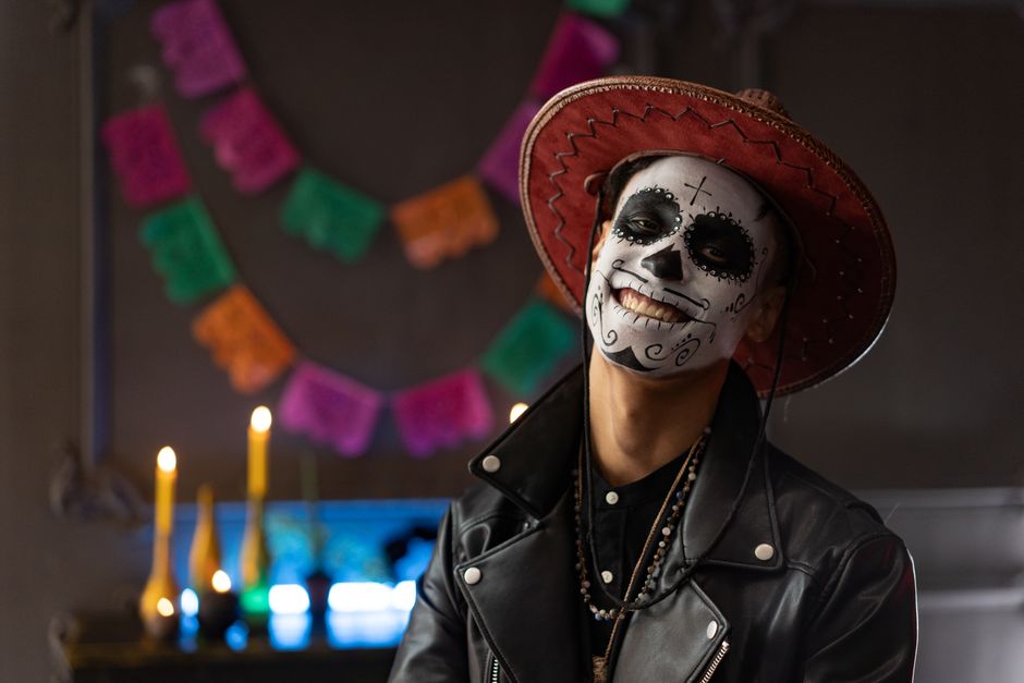 Man in Dia de los Muertos makeup smiling in traditional costume with decorations in the background.