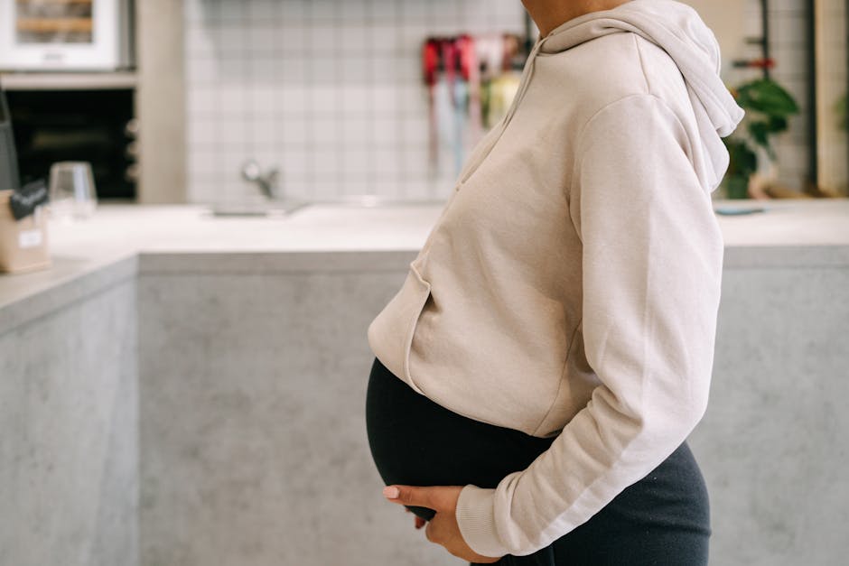 Pregnant woman in casual hoodie gently holding belly indoors.
