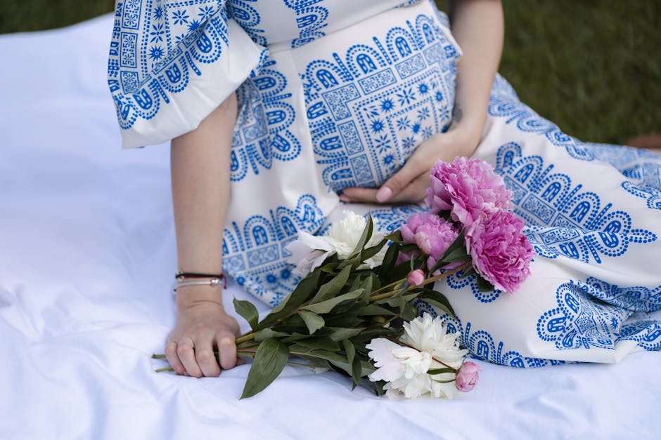 A serene scene of a pregnant woman in a blue patterned dress holding flowers outdoors.