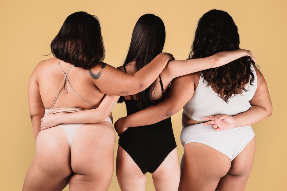 Back view of faceless female models in underwear hugging each other against beige wall in studio