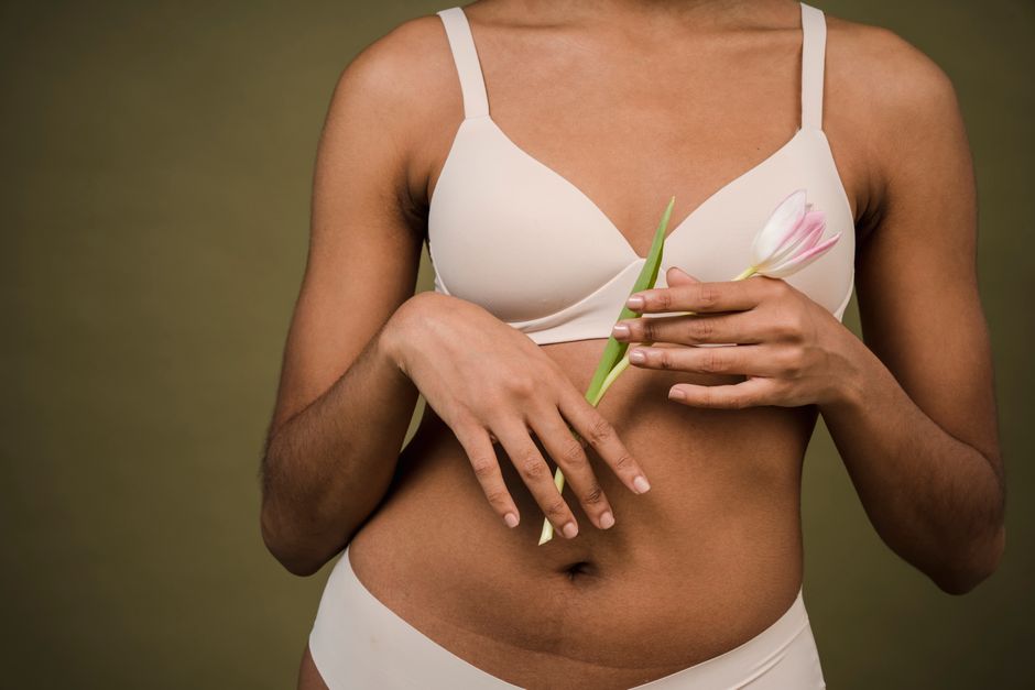 Crop unrecognizable African American female in white lingerie with hairs on arms standing against beige background with tulip in hands