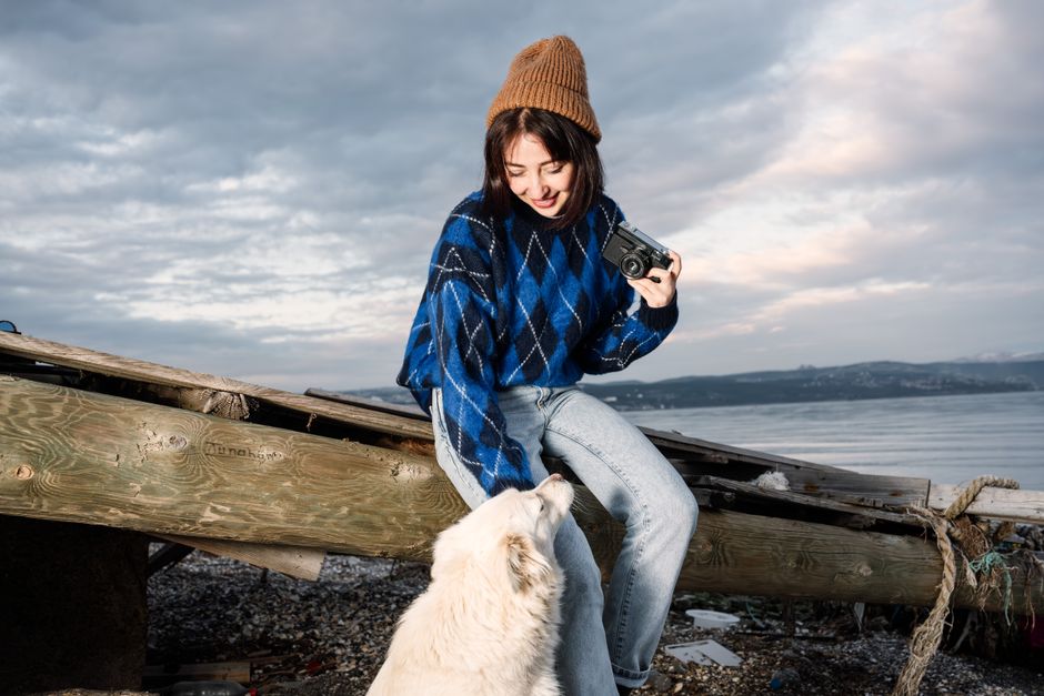 A young woman sitting by the sea with a camera, interacting with a white dog on a cloudy day.