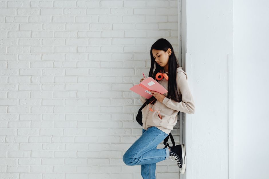 A young woman in casual attire writing in a notebook while leaning against a white brick wall.