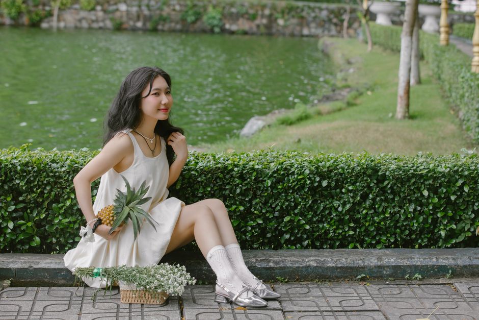 A young woman in a white dress sitting by a pond holding a pineapple, enjoying a serene outdoor setting.