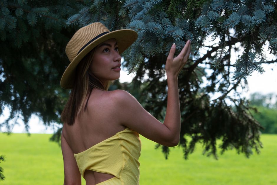 A woman in a straw hat touches an evergreen branch in a sunny outdoor setting.