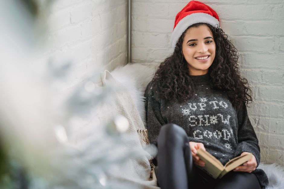 Young female in warm sweater sitting on floor and reading book while resting in cozy room on Christmas