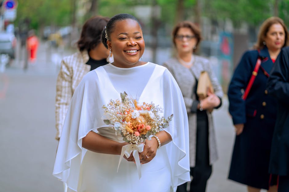 Smiling bride in white dress with bouquet surrounded by guests outdoors on a special day.