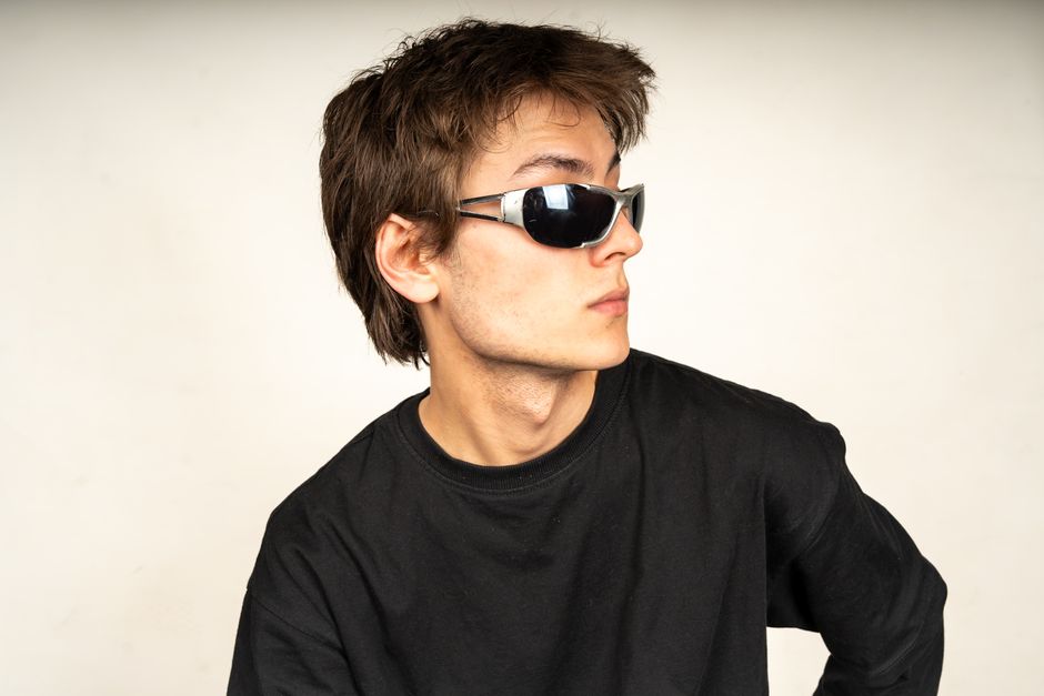 Portrait of a young man in a black shirt wearing sunglasses, gazing to the side against a plain background.