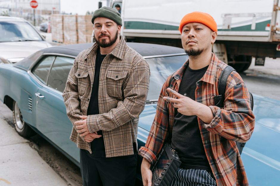 Two men in beanies pose confidently with a vintage car on an urban street, exuding style.
