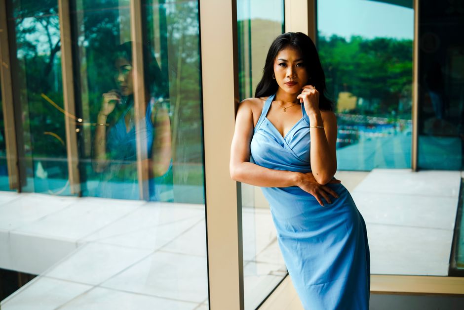 A stylish woman in a blue dress poses confidently by a large window with a reflection, capturing a modern and elegant look.